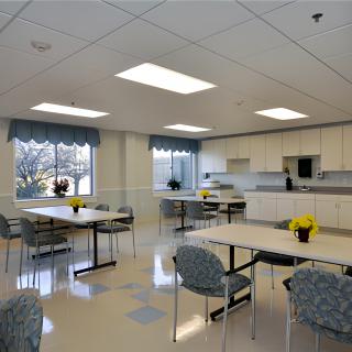 A spacious breakroom with multiple tables, chairs, and a kitchen area equipped with cabinets.