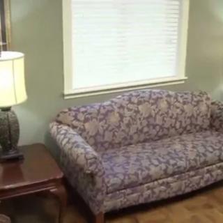 A cozy living room corner featuring a floral-patterned sofa, a wooden side table with a lamp, and a window with blinds.