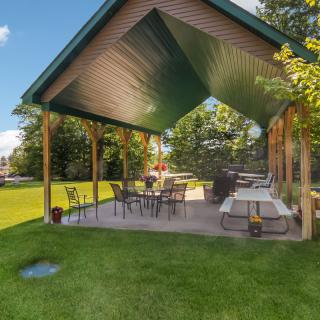 A cozy outdoor patio with a gazebo, furnished with tables and chairs, surrounded by a lush green lawn on a sunny day.