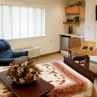 A cozy studio apartment with a stylish interior, featuring a bed, recliner chair, small kitchen area, and a decorative centerpiece on the table.