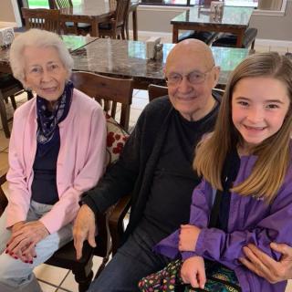 A young girl sits with her grandparents, all smiling warmly indoors.
