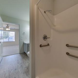 A modern bathroom with a shower featuring grab bars, leading to a well-lit hallway with wooden flooring.