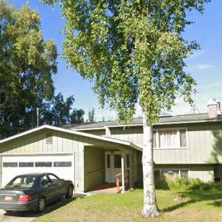 A suburban house with a garage and a car parked in the driveway, surrounded by trees on a sunny day.