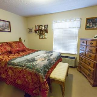 A cozy bedroom featuring a large bed with patterned bedding, wooden dresser, and artwork on the walls.