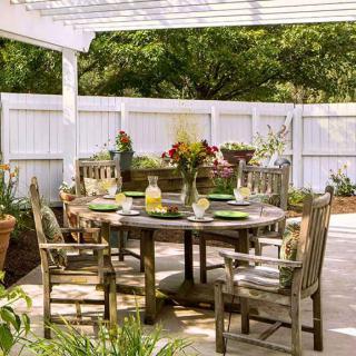 A cozy outdoor patio with a wooden dining set under a pergola, surrounded by lush greenery and potted plants.