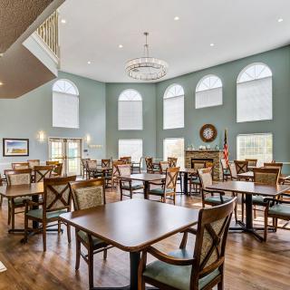 A spacious dining area with multiple tables and chairs, large windows, a chandelier, and wooden flooring.
