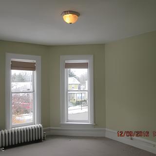 An empty room with carpet flooring, featuring two large windows, a radiator, and a ceiling light.