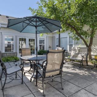 A cozy outdoor patio area featuring a table with an umbrella and chairs, set on a paved surface under bright sunlight, next to a tree.