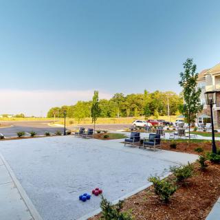 A sunny outdoor patio area with tables and chairs, surrounded by landscaped greenery and a distant view of trees.