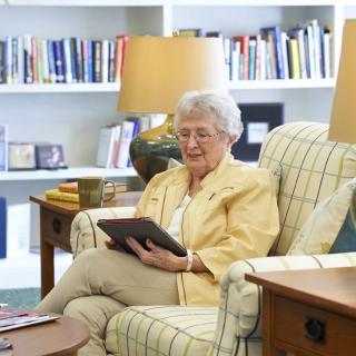 An elderly woman reading on a tablet in a cozy living room.