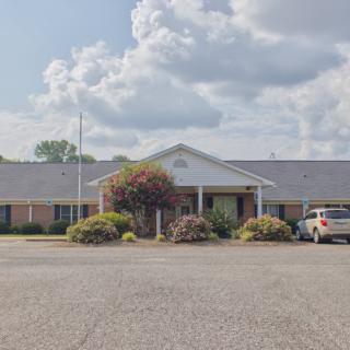 A single-story building with a parking lot in front, surrounded by trees under a partly cloudy sky.