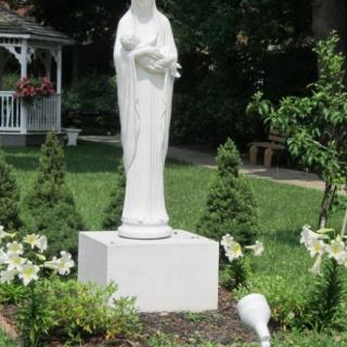 A religious statue stands in a beautifully landscaped garden surrounded by greenery and flowers.