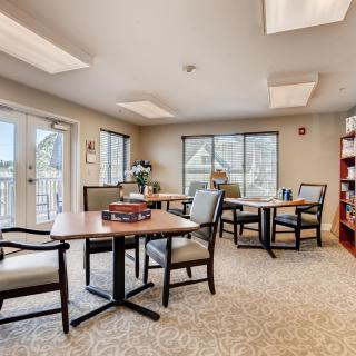 A cozy room with tables, chairs, and a bookshelf, featuring large windows and warm lighting.