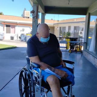 A person seated in a wheelchair outdoors, wearing a mask and appearing relaxed.