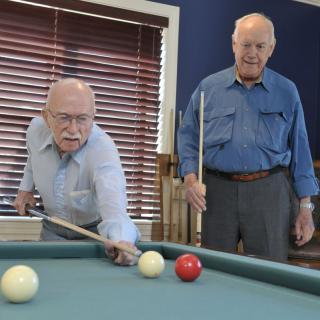 Two older men playing billiards in a room with blue walls and window blinds.