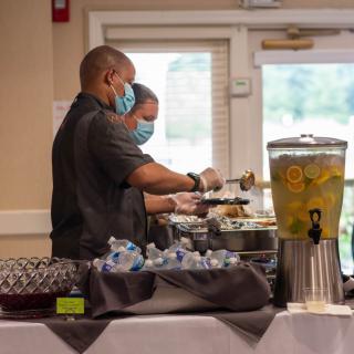 Caterers wearing masks serve food at a buffet during an indoor event.