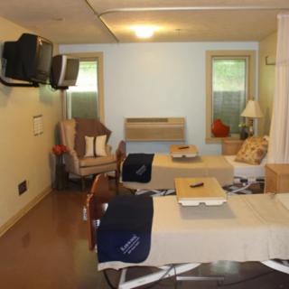 A room with two beds, a television, chairs, and windows letting in natural light.