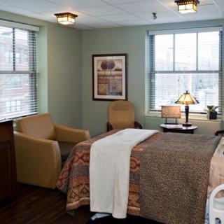 A cozy hospital room with a bed, comfortable chairs, and large windows letting in natural light.