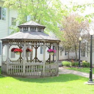 A wooden gazebo adorned with hanging flower baskets, surrounded by lush greenery and lampposts in a garden setting.