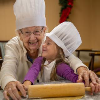 An elderly woman and a young girl wearing chef hats share a joyful moment while baking together.