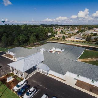 Aerial view of a building complex with a parking lot, surrounded by trees and suburban homes under a blue sky with scattered clouds.