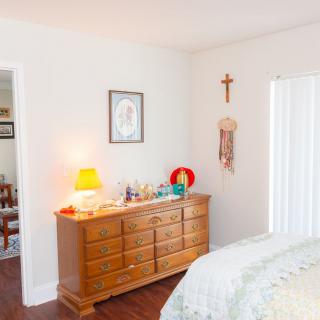 A cozy bedroom with a wooden dresser, illuminated by a small lamp and adorned with religious decor.