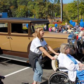 A person pushes another in a wheelchair past a vintage car at an outdoor event with blue balloons.
