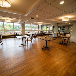 An empty dining room with tables and chairs, featuring large windows and wooden flooring.