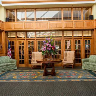 A well-furnished lobby with a central table adorned with flowers, surrounded by chairs and sofas, featuring wood paneling and large windows.