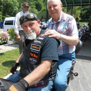 Two men sitting on a motorcycle outdoors, one wearing a leather vest and helmet, the other in casual clothing.