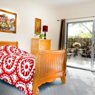 A cozy bedroom with wooden furniture, featuring a bed adorned with a red and white patterned duvet, artwork on the walls, and a sliding door leading outside.
