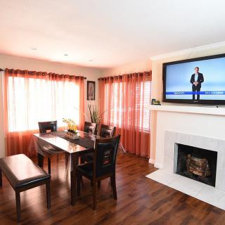 A cozy living and dining space with a fireplace, a television mounted above, wooden floors, and bright curtains.