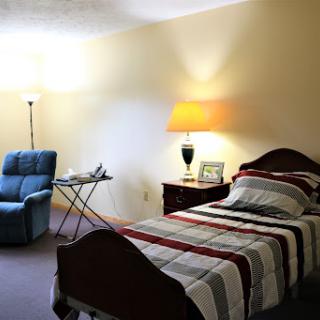 A cozy bedroom featuring a single bed with a striped blanket, an armchair, and a bedside lamp.