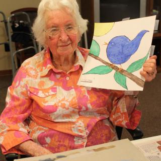 An elderly woman proudly displays her colorful drawing of a blue bird with leaves.