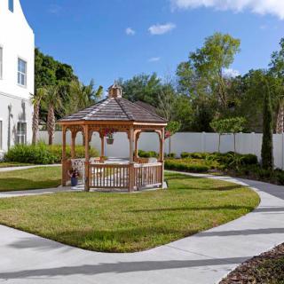 A picturesque gazebo in a garden setting with surrounding pathways, trees, and a nearby building.