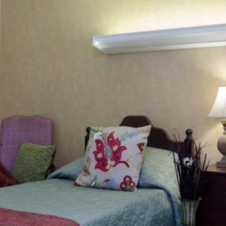 A cozy bedroom with a single bed adorned with colorful pillows, a bedside lamp, and a comfortable chair in the corner.