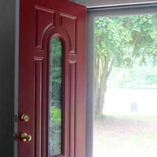 A red door with a glass panel stands open, allowing sunlight to filter through and revealing a view of an outdoor tree.