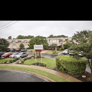 An apartment complex with a parking lot and several parked cars, surrounded by greenery under an overcast sky.