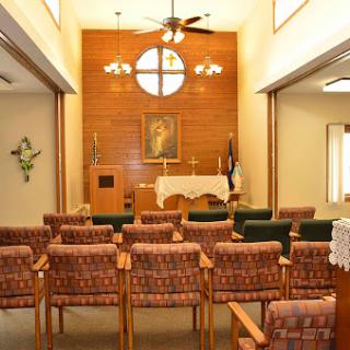 A serene chapel interior featuring rows of chairs facing an altar, adorned with wooden paneling and a large window.