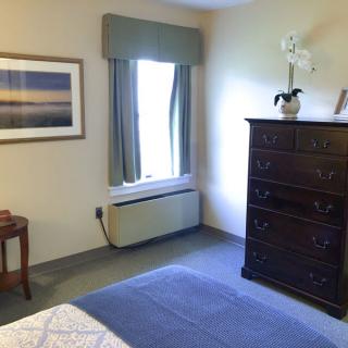 A cozy bedroom with a tall dresser, a window with curtains, and a painting on the wall.