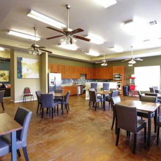 A spacious dining area with multiple tables and chairs, featuring a kitchen area and ceiling fans.