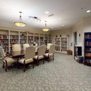 A cozy library room with a large table surrounded by chairs, filled with shelves of books and soft lighting.
