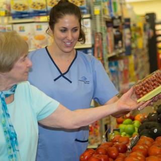 An elderly woman receives assistance from a caregiver while shopping for produce in a grocery store.