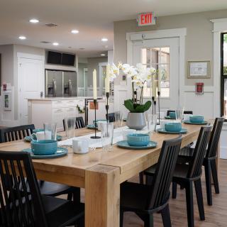 A modern dining room with a wooden table elegantly set with teal dishes and an orchid centerpiece, adjacent to an open kitchen.