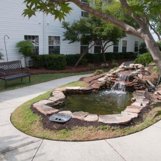 A peaceful garden scene with a stone-lined pond and waterfall, featuring a bench beside a pathway.
