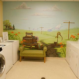 A cozy laundry room featuring a mural of a rural landscape with washing machines and a green couch.