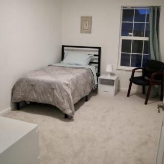A minimalist bedroom with a single bed, nightstand, lamp, and chair by the window.