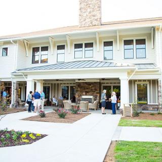 A group of people gathering outside a large house with a spacious patio and well-kept lawn.