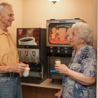 Two elderly individuals enjoy coffee and conversation by a beverage machine.