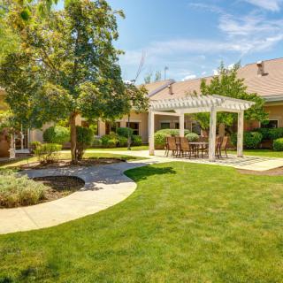 A beautifully landscaped backyard featuring a pergola with outdoor seating, surrounded by lush grass and trees on a sunny day.